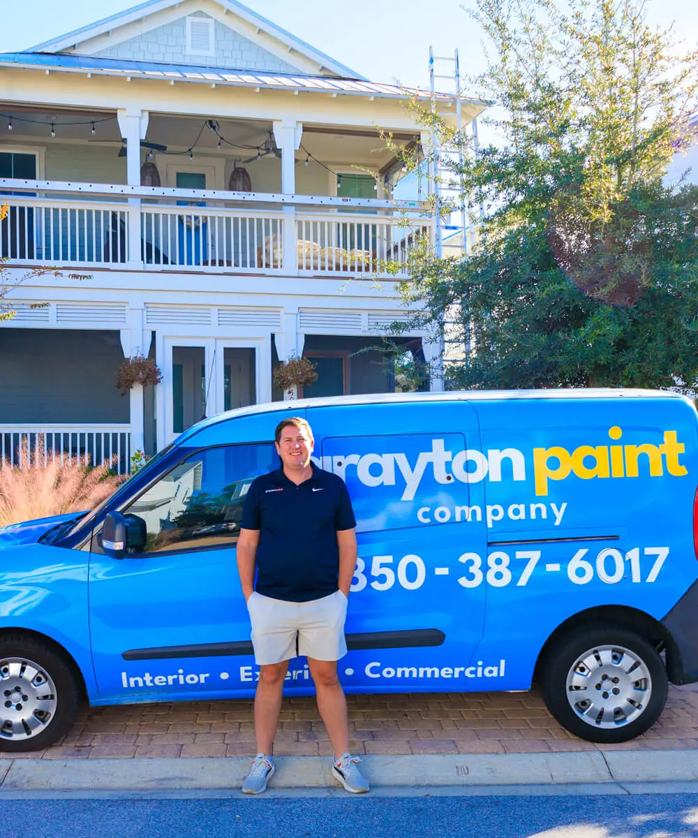 A professional portrait of the owner standing in front of a blue Grayton Painting company van, parked before a beautiful two-story coastal home with freshly painted white trim.